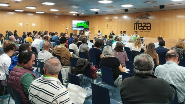 A conference room filled with attendees seated in rows, actively listening to a panel discussion. The room features wooden walls with the logo "IFEZA" prominently displayed, indicating the venue. A screen at the front shows visual content, and the panelists are seated at a long table, engaging in discussions about the event's theme. The setting suggests a professional forum or seminar.