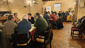 A group of participants seated at tables in a cozy, rustic room with warm lighting and wooden furniture. The attendees are engaged in discussions, collaborating in small groups. A roll-up banner and a projection screen are visible in the background, indicating an organized workshop or seminar setting. Coats and bags are placed on chairs and a rack along the wall.