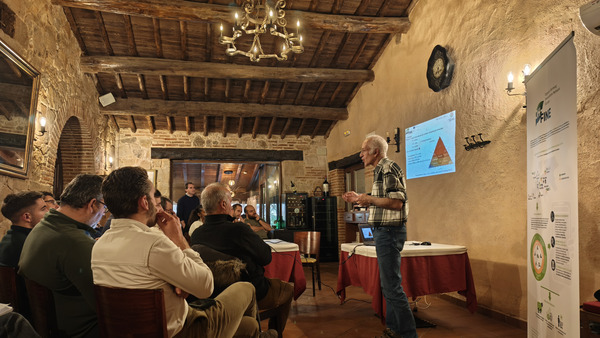 A rustic meeting room with exposed wooden beams and stone walls. A presenter is standing near a projection screen displaying a presentation slide with a pyramid diagram. A roll-up banner with informational content is positioned to the right. Attendees are seated at tables, attentively listening to the presentation. The setting is warm and inviting, with a chandelier hanging from the ceiling and soft lighting.