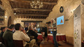 A rustic meeting room with exposed wooden beams and stone walls. A presenter is standing near a projection screen displaying a presentation slide with a pyramid diagram. A roll-up banner with informational content is positioned to the right. Attendees are seated at tables, attentively listening to the presentation. The setting is warm and inviting, with a chandelier hanging from the ceiling and soft lighting.